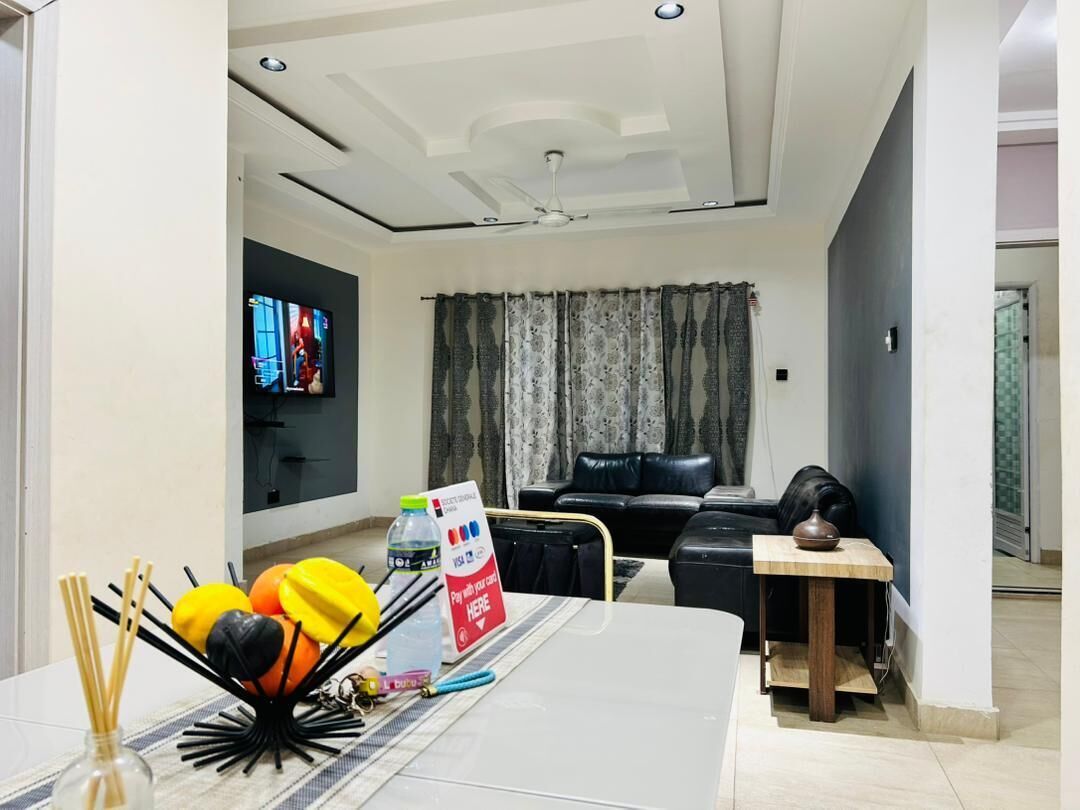A living room with a TV, a black leather sofa, a dining table with a vase, and a ceiling fan.
