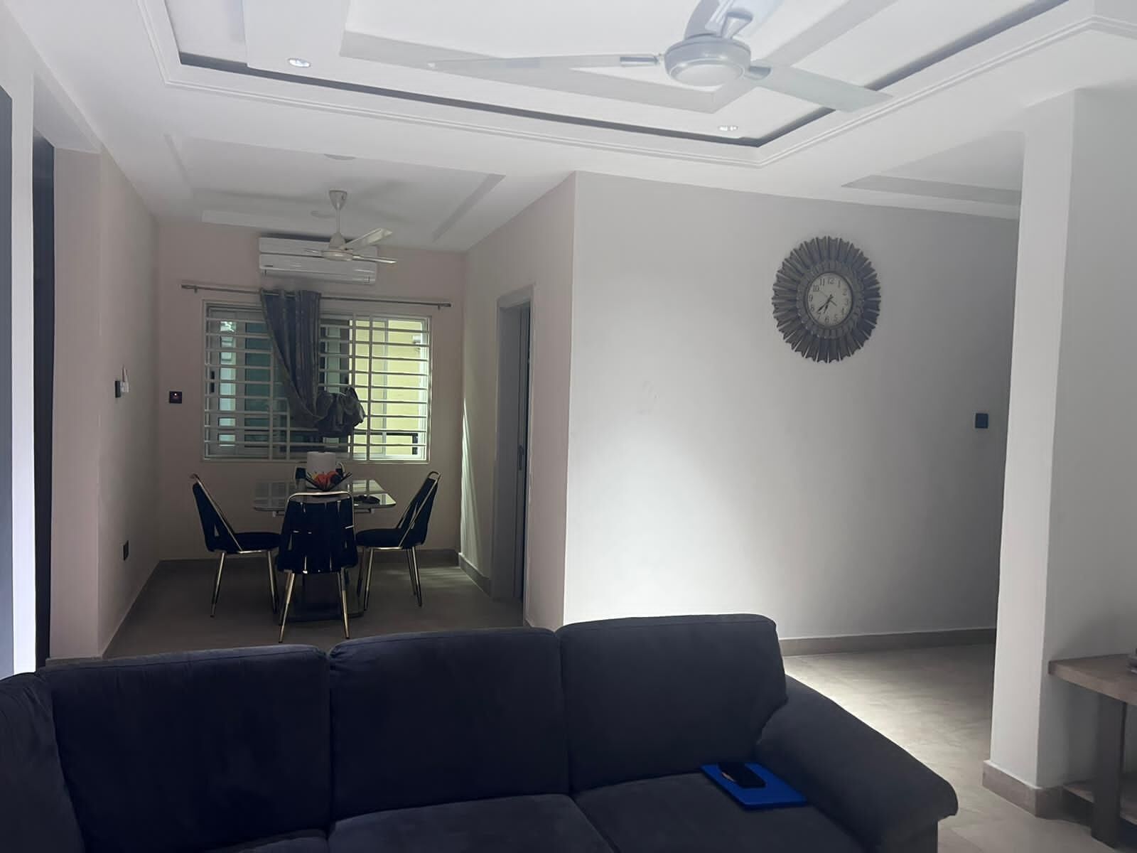 Uma sala de estar com um sofá escuro, um espaço de jantar com mesa e cadeiras, e um ventilador de teto.