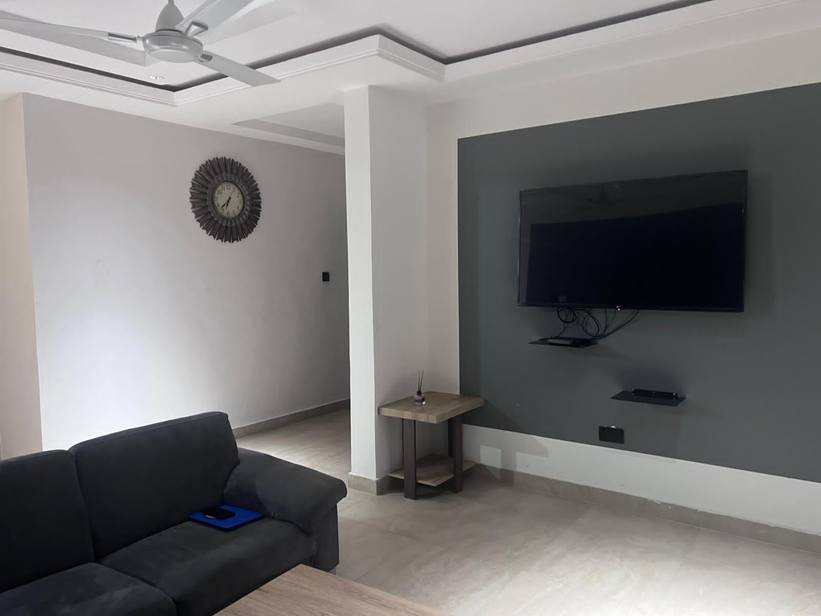 Ein modernes Wohnzimmer mit einem dunklen Sofa, einem kleinen Tisch, einem Wand-Fernseher und einem Deckenventilator.