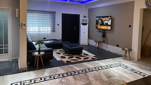 A modern living room with a sofa, a coffee table, a TV mounted on the wall, and a ceiling fan. - T-Roys Apartments (Accra)