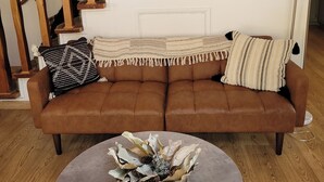 A brown leather sofa with a woven throw blanket and two pillows, a round coffee table with a pinecone centerpiece, and a staircase with wooden railings. - Charming 2-bdrm cabin in Fawnskin, with Wifi & just a 5 minute walk to the lake (Fawnskin)
