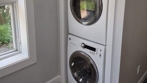 Lavadora y secadora apiladas, blancas y compactas, en una habitación con ventana y una pequeña planta en la parte superior.