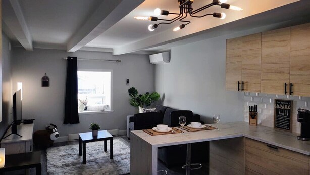 A modern kitchen with a dining area, a TV, and a cozy seating space with a plant. - Panache Royale 2.... (Québec City)