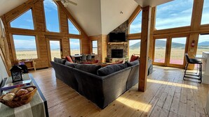 Un salón amplio con un sofá modular gris grande, una chimenea de piedra y un televisor de pantalla plana. Grandes ventanas ofrecen vistas amplias del paisaje circundante.