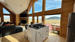Una sala de estar amplia con chimenea de piedra, una cocina con estufa a gas y grandes ventanales con vistas al paisaje desértico.