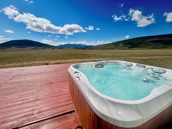 Ein Whirlpool auf einer Holzterrasse mit herrlicher Aussicht auf eine Wiese und Berge im Hintergrund.