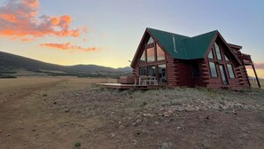 Una cabaña de troncos con techo verde, ventanas grandes y una terraza, ubicada en un paisaje árido y abierto con colinas al fondo.