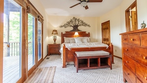 Un dormitorio con cama de madera, mesita de noche con lámpara, ventilador de techo, cómoda y un gran ventanal con vistas a zonas verdes.