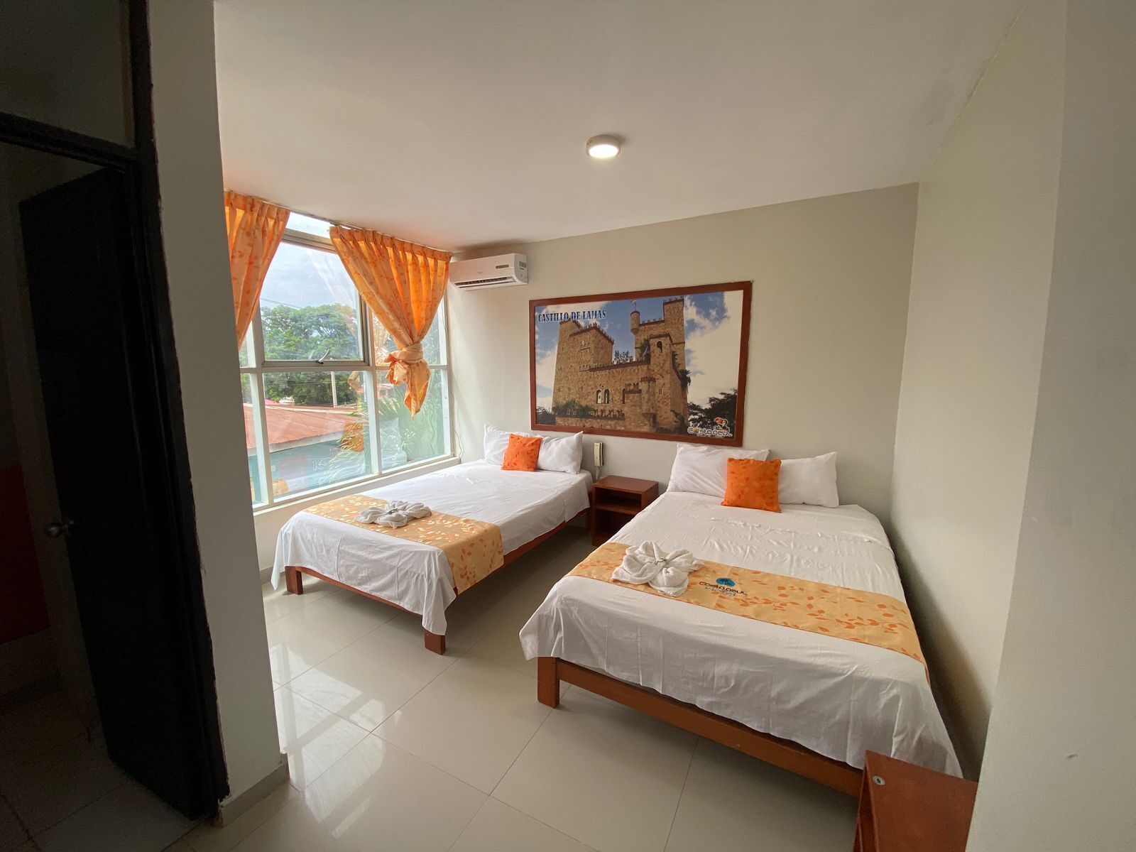A hotel room with two beds, a window with curtains, an air conditioning unit, and a painting of a castle.