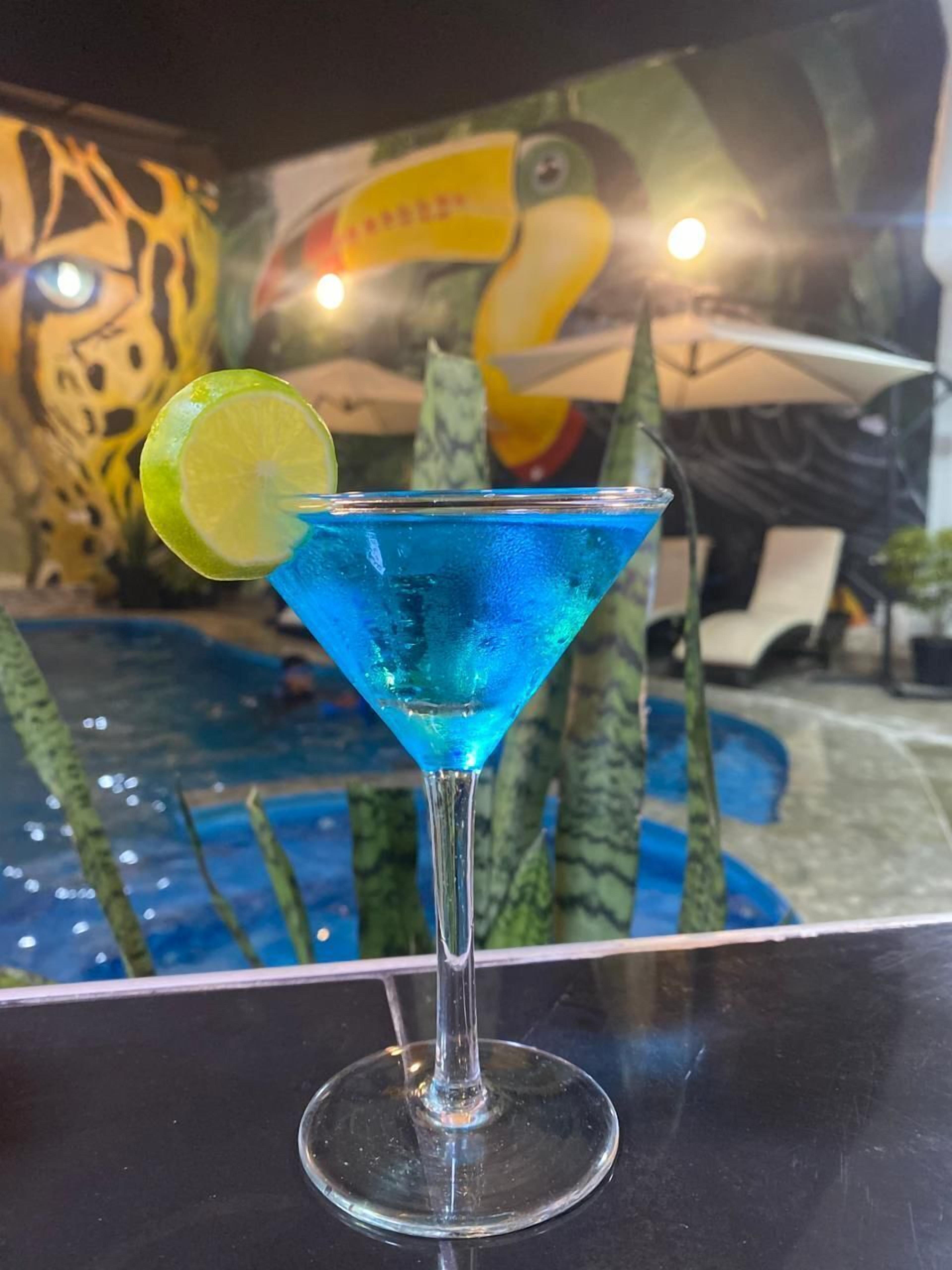 Cóctel azul con un adorno de lima en una copa de martini, sobre una superficie reflectante.