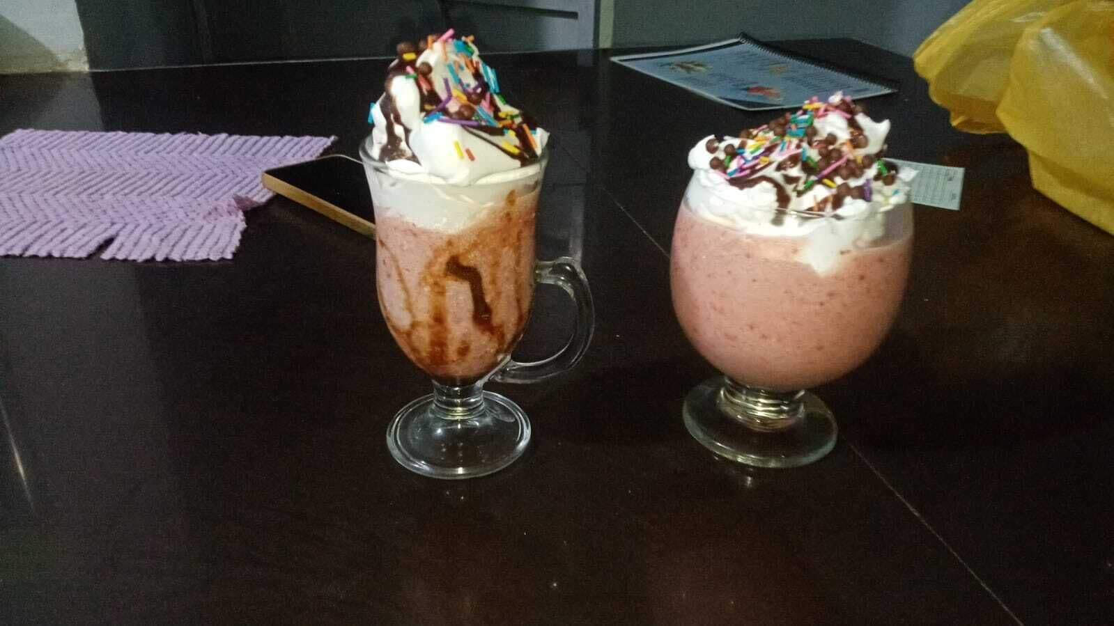 Two milkshakes in glasses with whipped cream and chocolate syrup on a dark countertop.