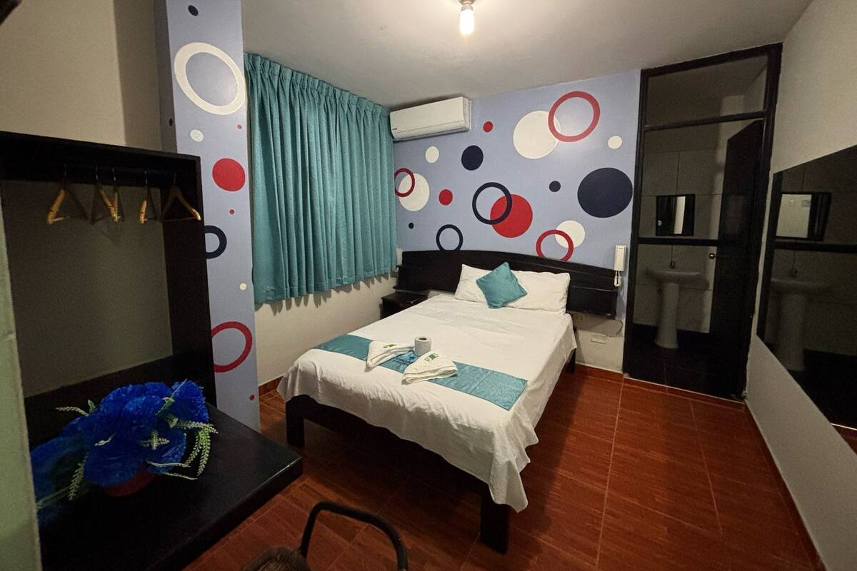 Une chambre d’hôtel comprenant un lit, un bureau, un miroir, une armoire, une salle de bain avec un lavabo et des toilettes, et un mur orné de motifs circulaires.