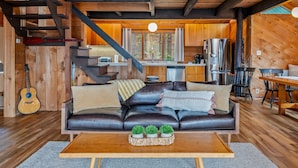 A cozy cabin interior with a wooden staircase, a dining area, a kitchen, and a living room with a leather sofa and a coffee table.