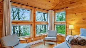 A cozy cabin room with wooden walls, large windows, a bed, a chair, and a lamp.