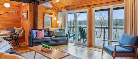 Ein gemütliches Blockhaus-Interieur mit Holzbalkendecke, einer Couch, einem Esstisch und einer großen Schiebeglas-Tür, die auf eine Außenterrasse führt.