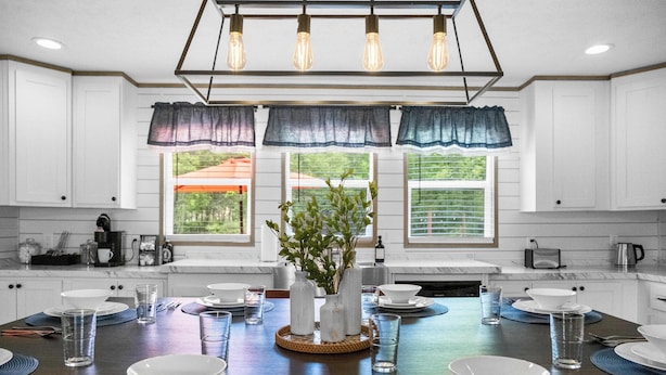 Uma cozinha moderna com armários brancos, uma mesa de jantar de madeira posta com pratos, tigelas e copos brancos, e uma planta no centro.