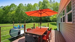 Um deck de madeira com mesa, cadeiras, um guarda-sol vermelho e uma churrasqueira.