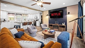 Uma sala de estar moderna com sofá, poltronas e mesa de centro. Há um ventilador de teto e uma TV de tela plana.