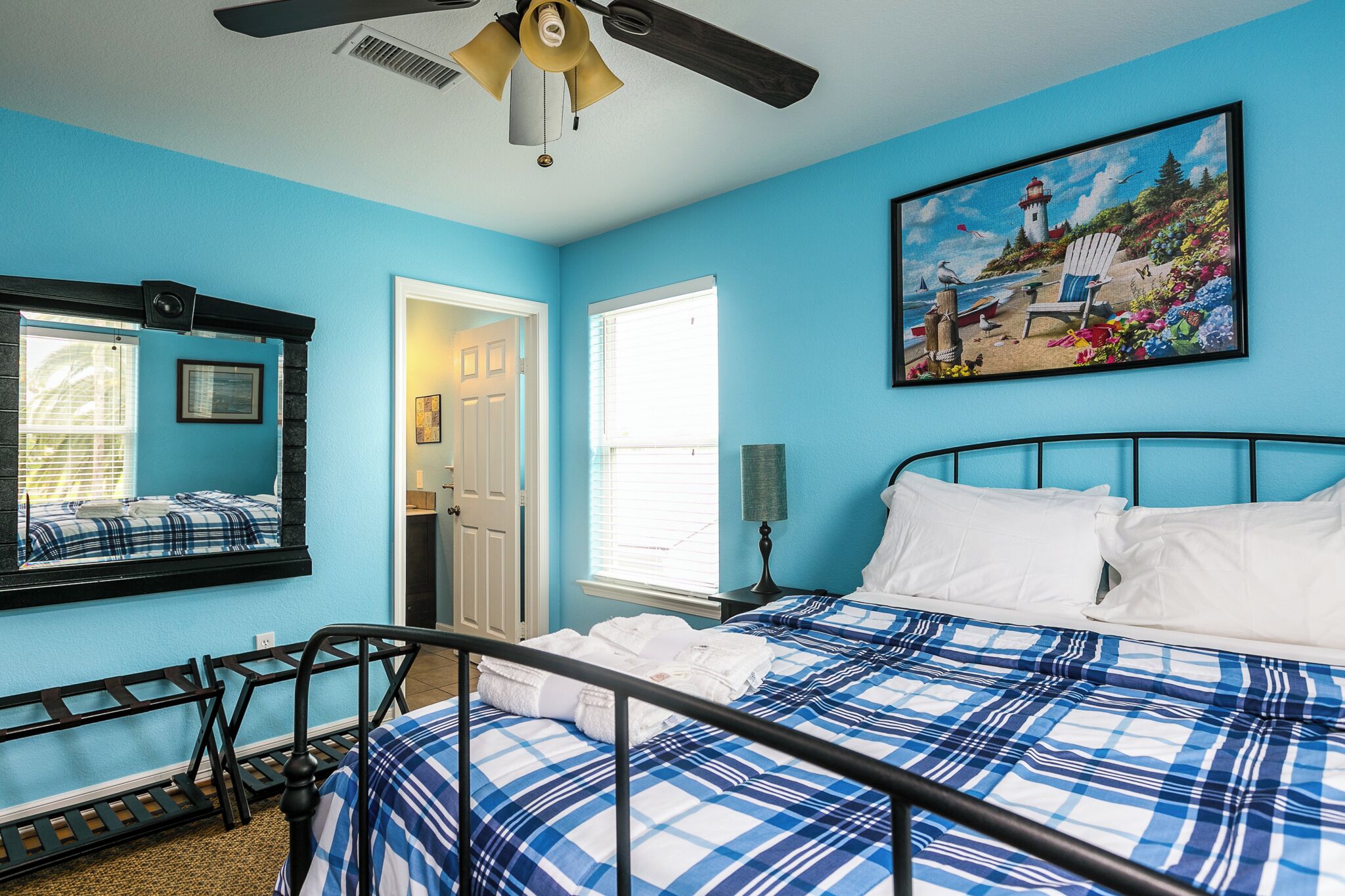 Une chambre à coucher avec un lit, un ventilateur de plafond, un tableau représentant un phare et une fenêtre avec des stores.