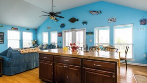 Una cocina con pared azul, armarios de madera, una zona de comedor con sillas y un ventilador de techo.