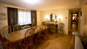 Een eetkamer met een houten tafel en stoelen, een raam met gordijnen, een spiegel boven de tafel, en een gang die naar een andere kamer leidt.