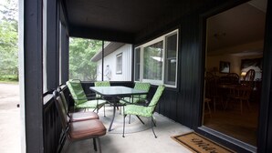 Een overdekt terras met een tafel en stoelen, een welkomstmat en uitzicht op het huis door grote ramen.