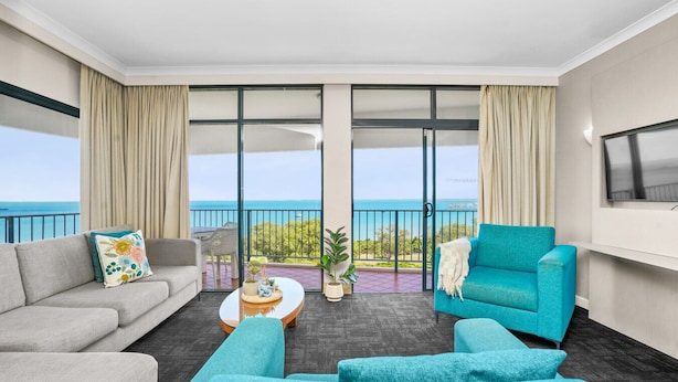 Habitación de hotel con balcón, un sofá gris, un sillón azul, una mesa de centro y un televisor montado en la pared.