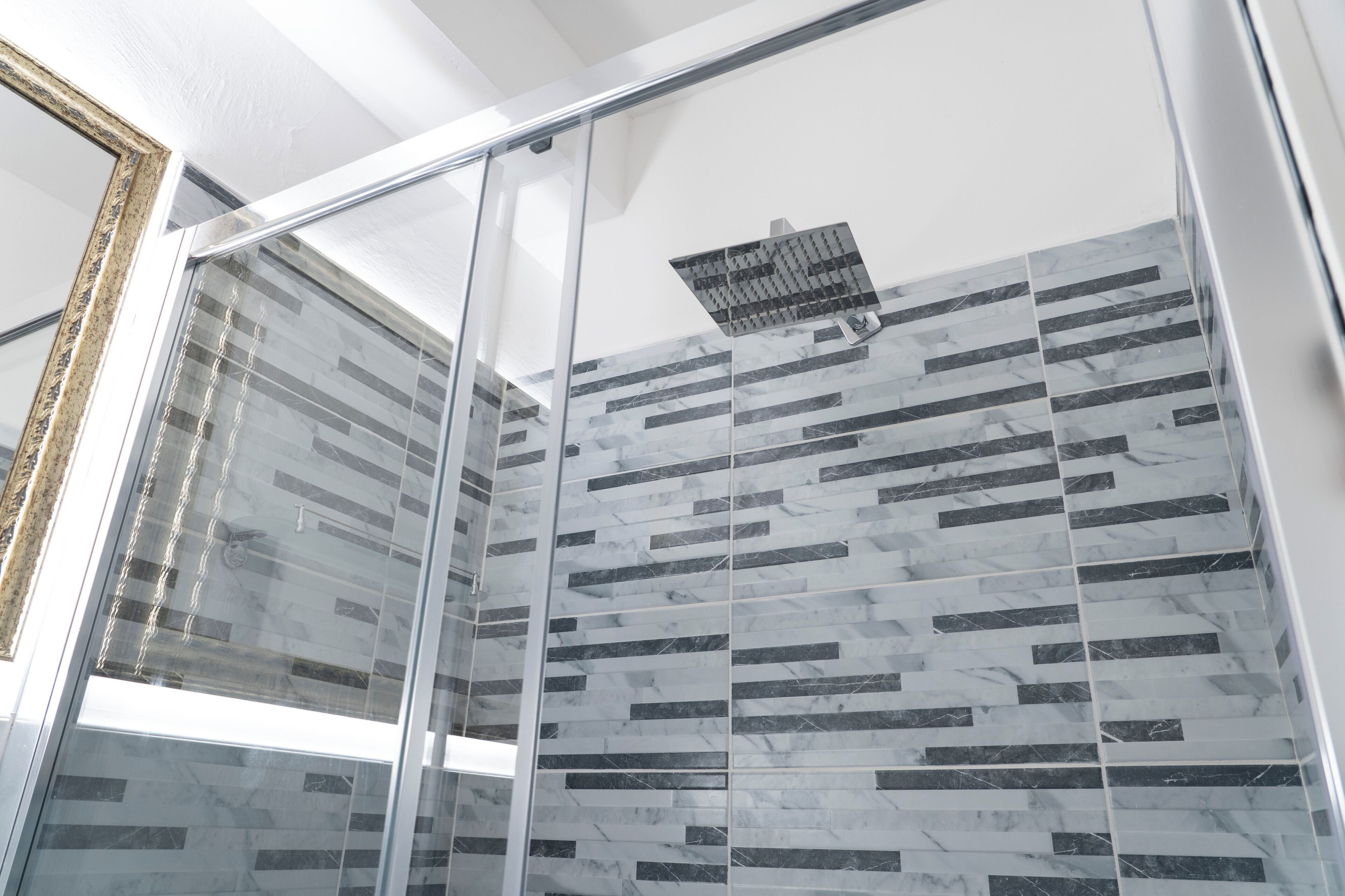 A modern bathroom with a glass shower enclosure, a tiled wall with a geometric pattern, and a visible ceiling vent.