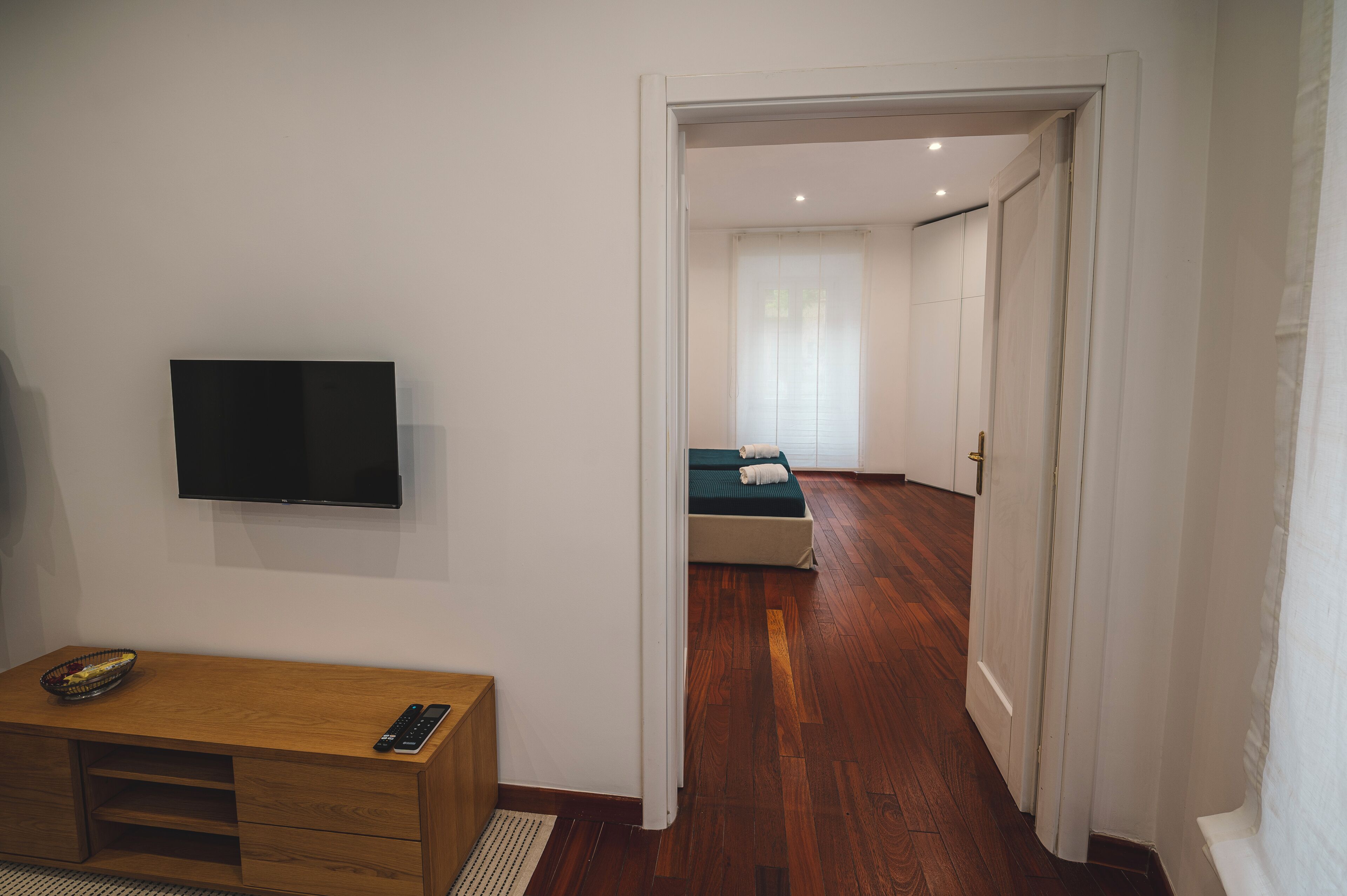 Una stanza con una scrivania in legno, una televisione a schermo piatto, un letto con materasso verde e una porta bianca che dà su un'altra stanza.