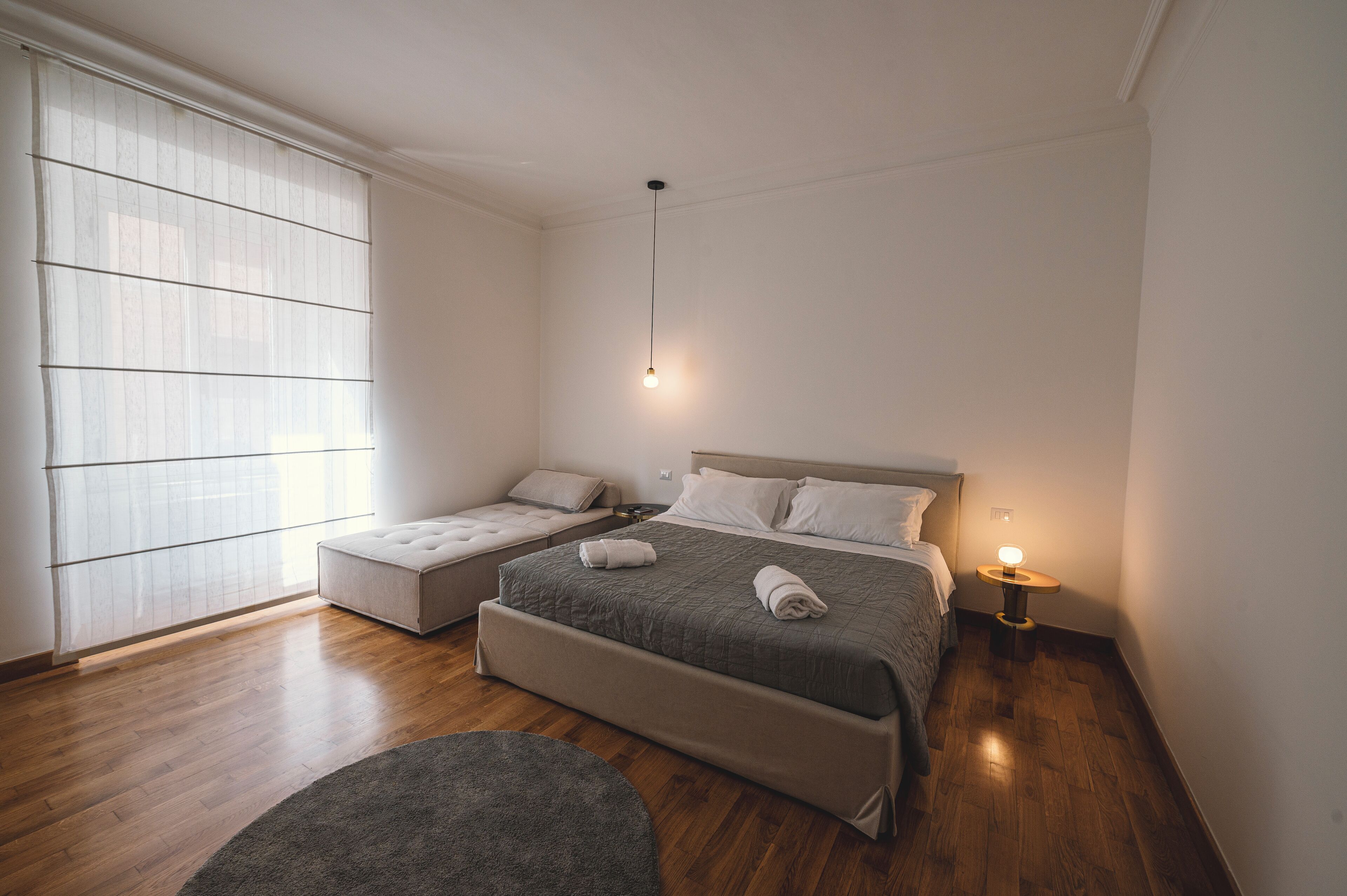 Una camera da letto moderna con un letto grande, un divano letto più piccolo, un comodino con una lampada e un'ampia finestra con tende trasparenti.