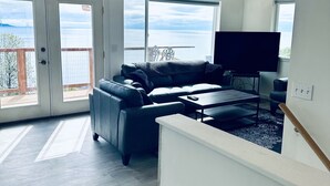 A spacious living room with a ceiling fan, a large window with a view of the ocean, a dark-colored sofa, and a flat-screen TV on a stand. - Paradise Suites Eagles Nest Apt C (Homer)