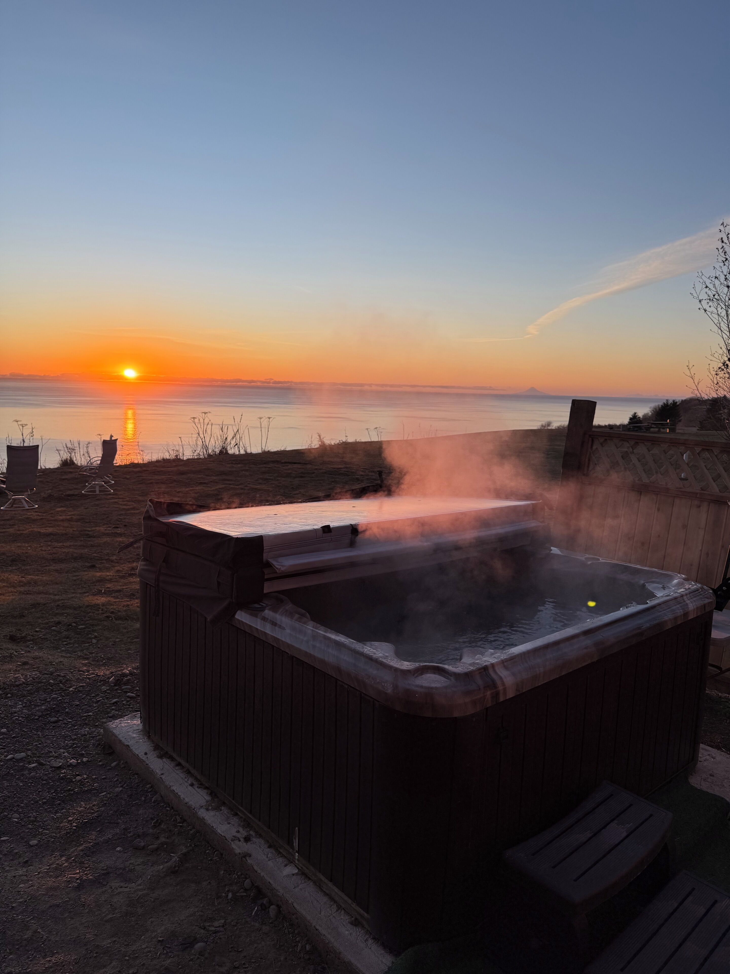 Un bain à remous doté d’une terrasse en bois, entouré d’une clôture en bois, avec vue sur une étendue d’eau au coucher du soleil.