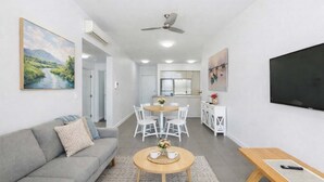 A modern living room with a grey sofa, a coffee table, a dining area with a white table and chairs, a kitchen with white cabinets, and a flat-screen TV mounted on the wall. - Holborn 2BR Apartment with Pool, Gym & Parking in Townsville CBD (Townsville City)