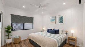 Un dormitorio con cama, ventilador de techo, ventana con persianas, aire acondicionado instalado en la pared y una mesita de noche con lámpara.
