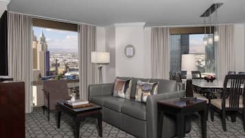 Een moderne hotelkamer met een grijze leren bank, een klein bijzettafeltje, een eethoek met tafel en stoelen, en grote ramen met uitzicht op de stad.