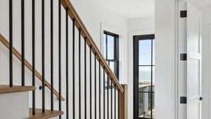 A modern staircase with wooden steps and black balusters, leading to a room with large windows and a view of the sea. - The Lookout | Private Pool | Steps to Beach & Pier | Newly Built (Folly Beach)