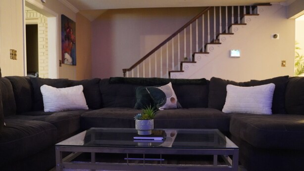 A living room with a black leather sofa, a glass coffee table with a plant, and a staircase in the background.