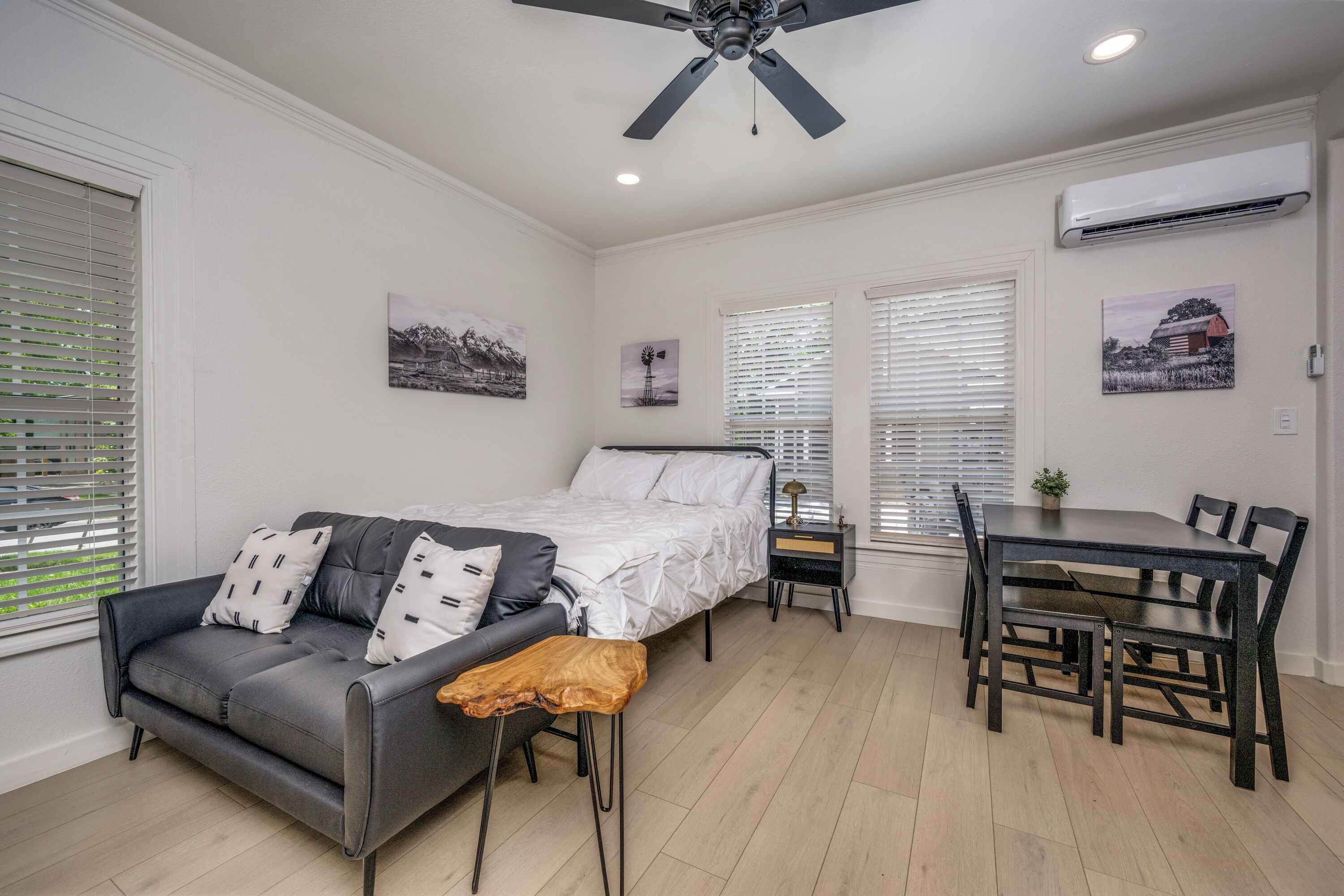A bedroom with a bed, a gray sofa, a wooden bench, a dining table with chairs, and a ceiling fan.