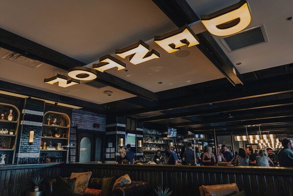 A bar area with a modern design, featuring a prominent illuminated sign, shelves with bottles, and a seating area with couches. - The Vanderbilt (Denton)