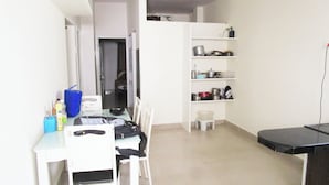 A compact kitchen with white cabinetry, a dining table, and chairs. There is a small stool and a shelf with kitchenware.