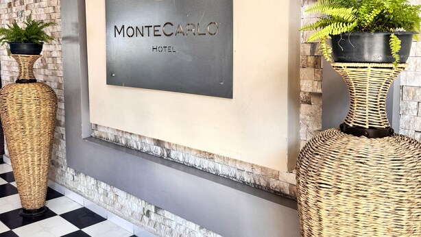 Uma placa emoldurada com o logotipo e o nome do Hotel Monte Carlo, ladeada por dois grandes vasos de vime com plantas.