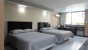 Quarto de hotel com duas camas, uma mesinha com garrafas de água, janela com cortinas e aparelho de ar condicionado.
