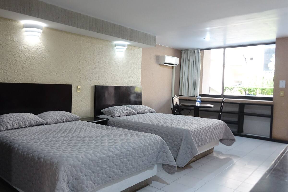 Ein Hotelzimmer mit zwei Betten, einem kleinen Tisch mit Wasserflaschen, einem Fenster mit Vorhängen und einer Klimaanlage.