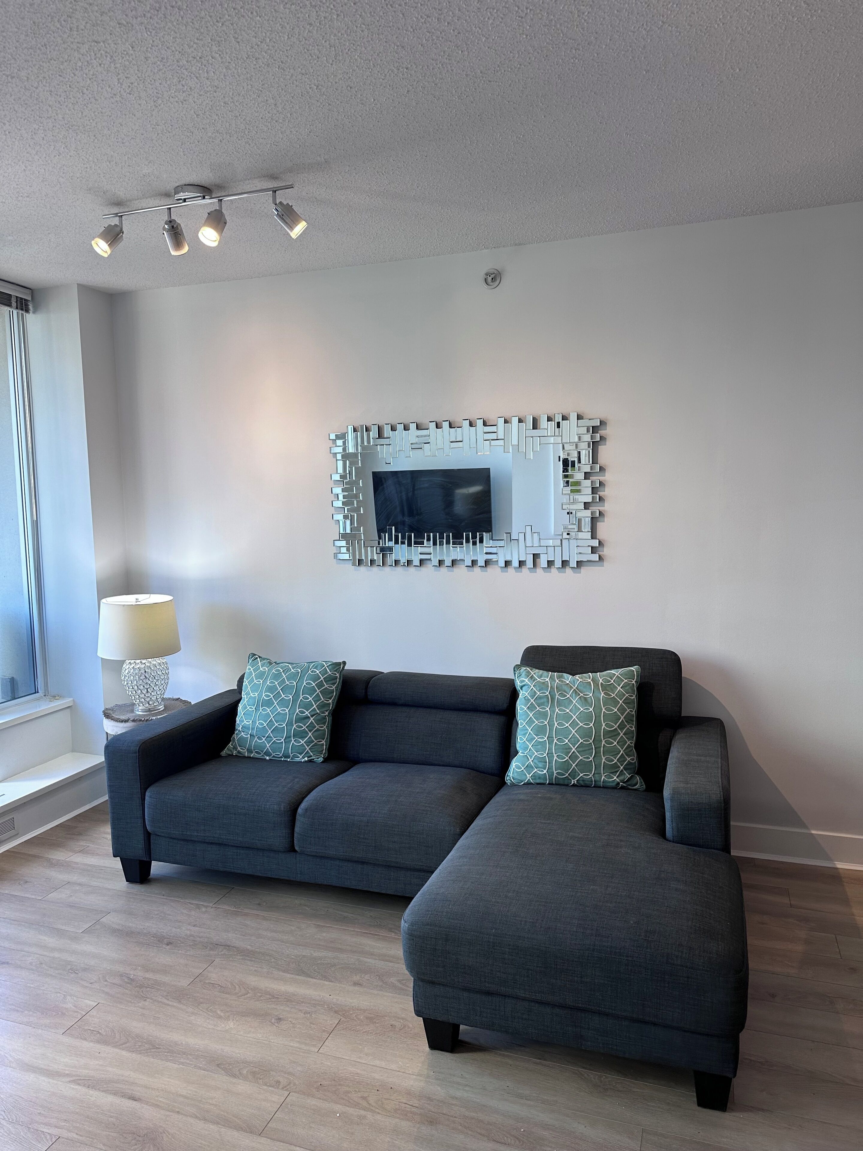 A modern living room with a dark grey sectional sofa, a matching ottoman, a decorative wall mirror, and a floor lamp.