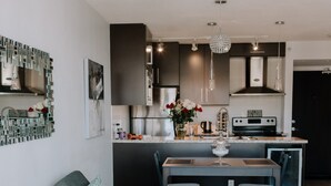 Moderni olohuone, jossa on harmaa kulmasohva, ruokapöytä ja taustalla keittiötila.