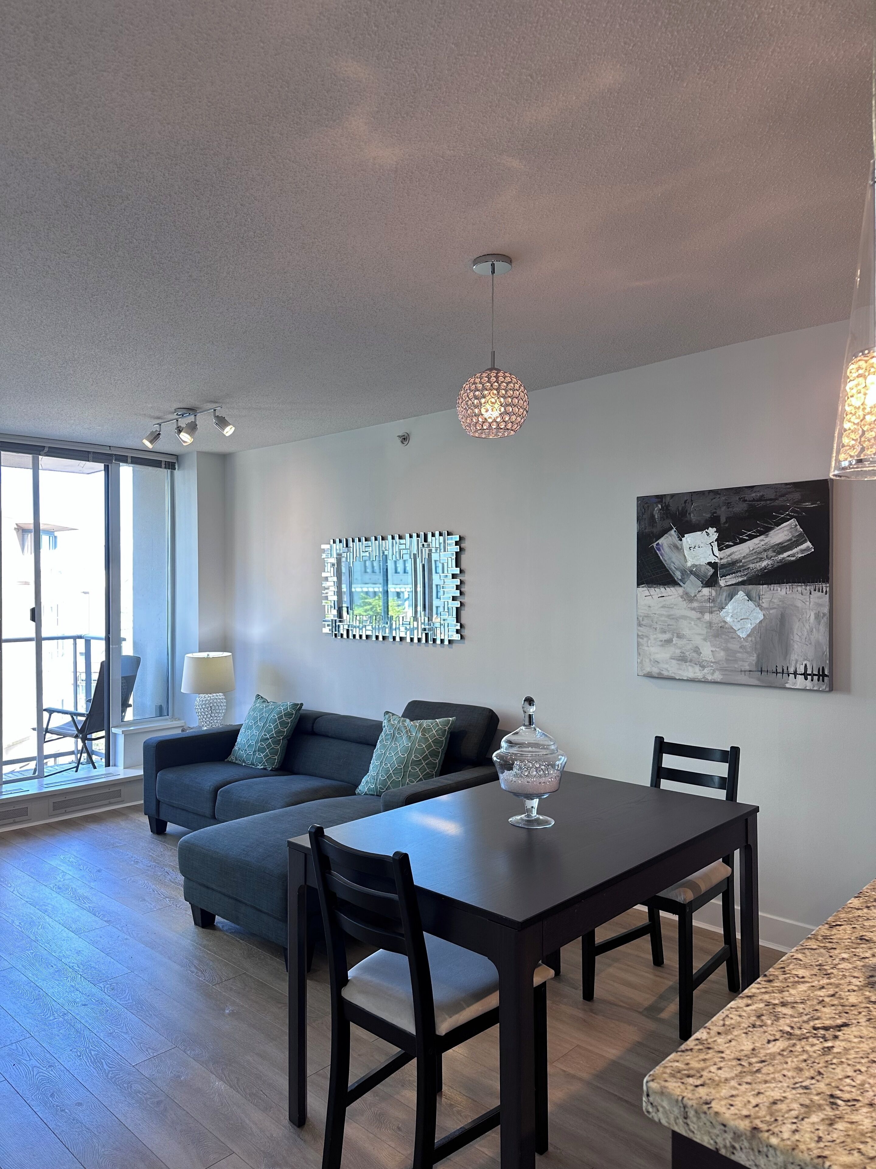 A modern living room with a dark sofa, a dining table, and chairs. There are abstract wall art pieces and a large window providing natural light.
