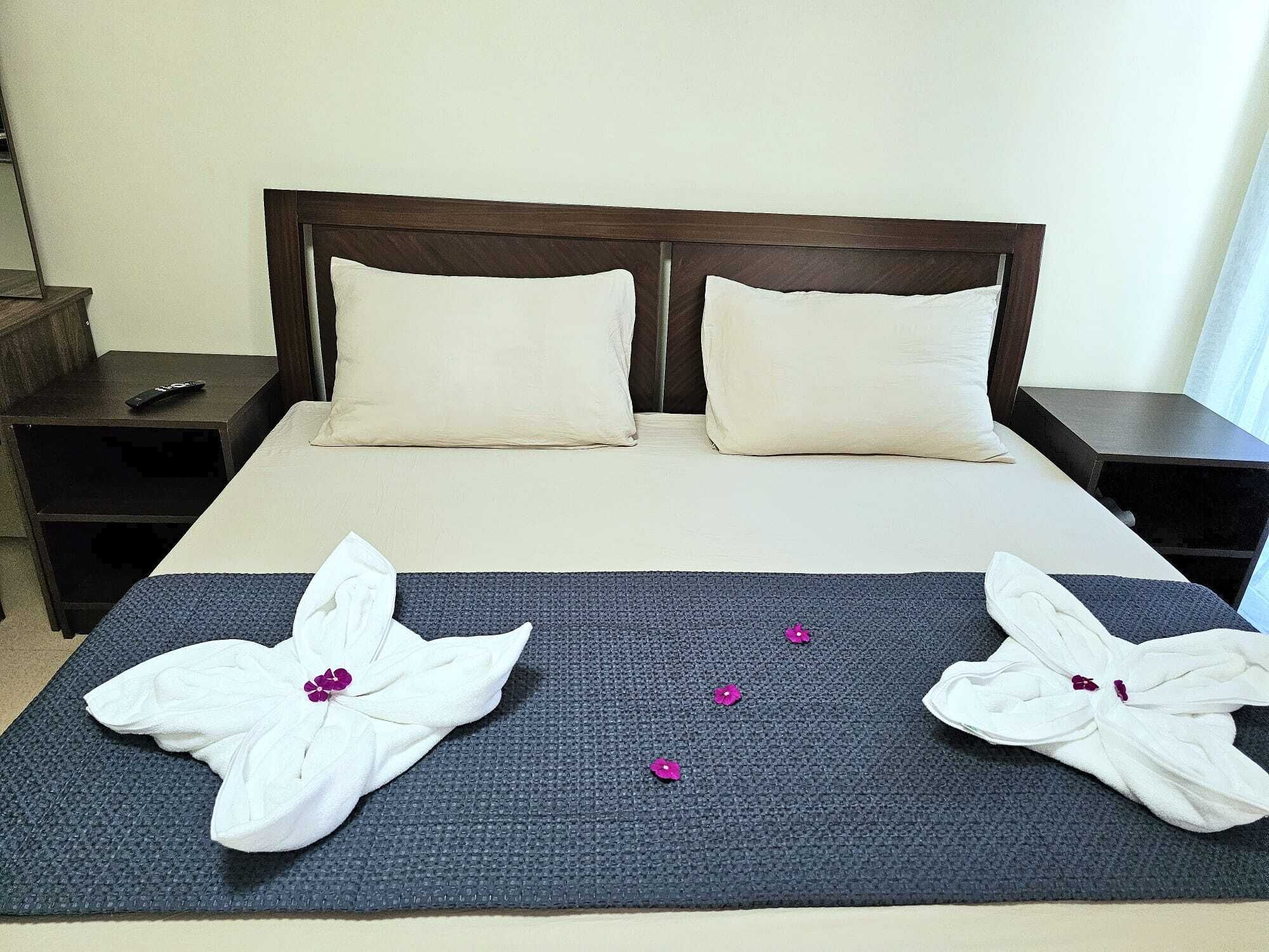 A neatly made bed with two pillows, a dark blue bedspread, and two towels folded into flower shapes with purple flowers.