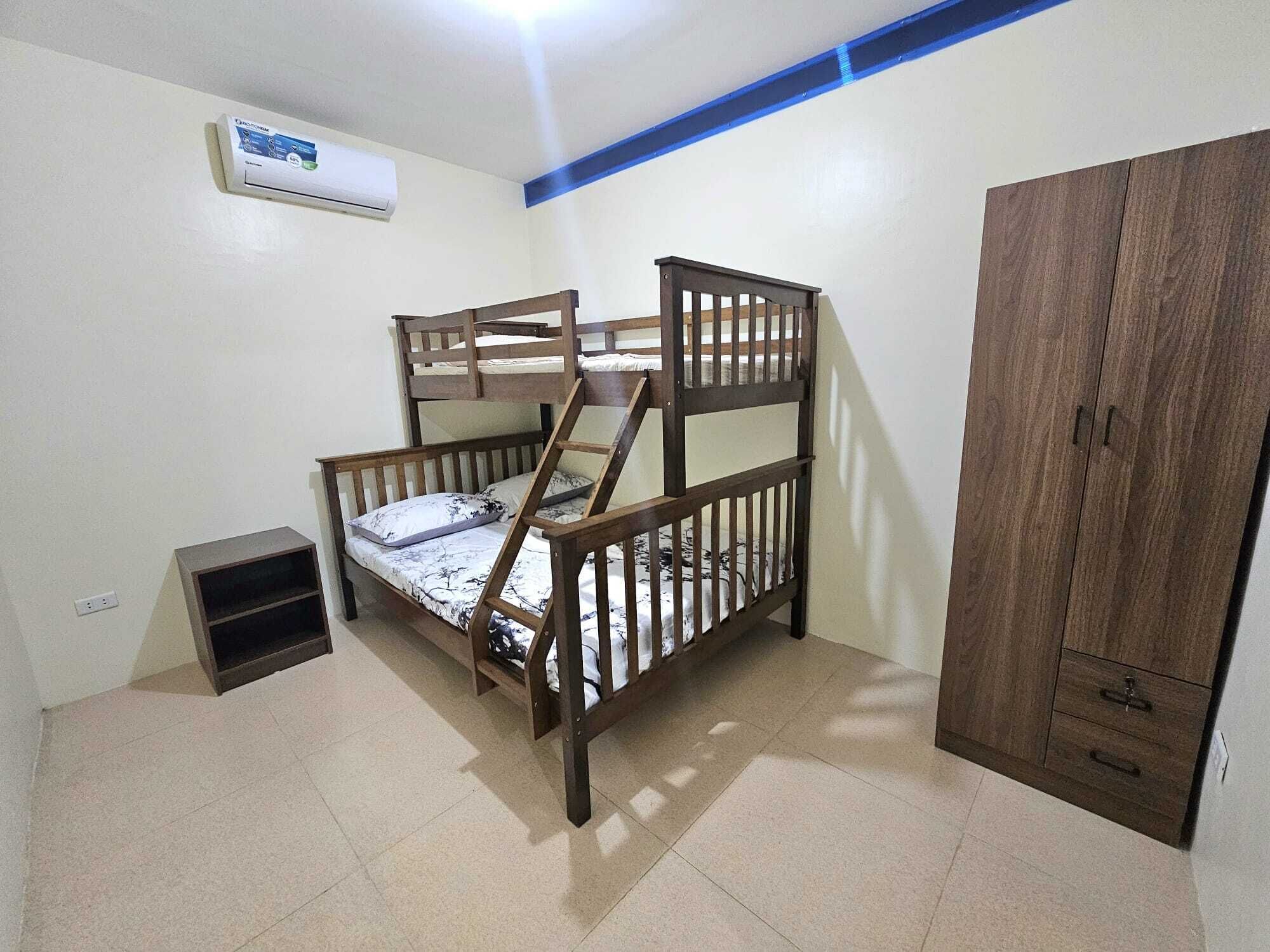 A bunk bed with a wooden frame and a small shelf below it, a wardrobe with two drawers, and an air conditioning unit on the wall.