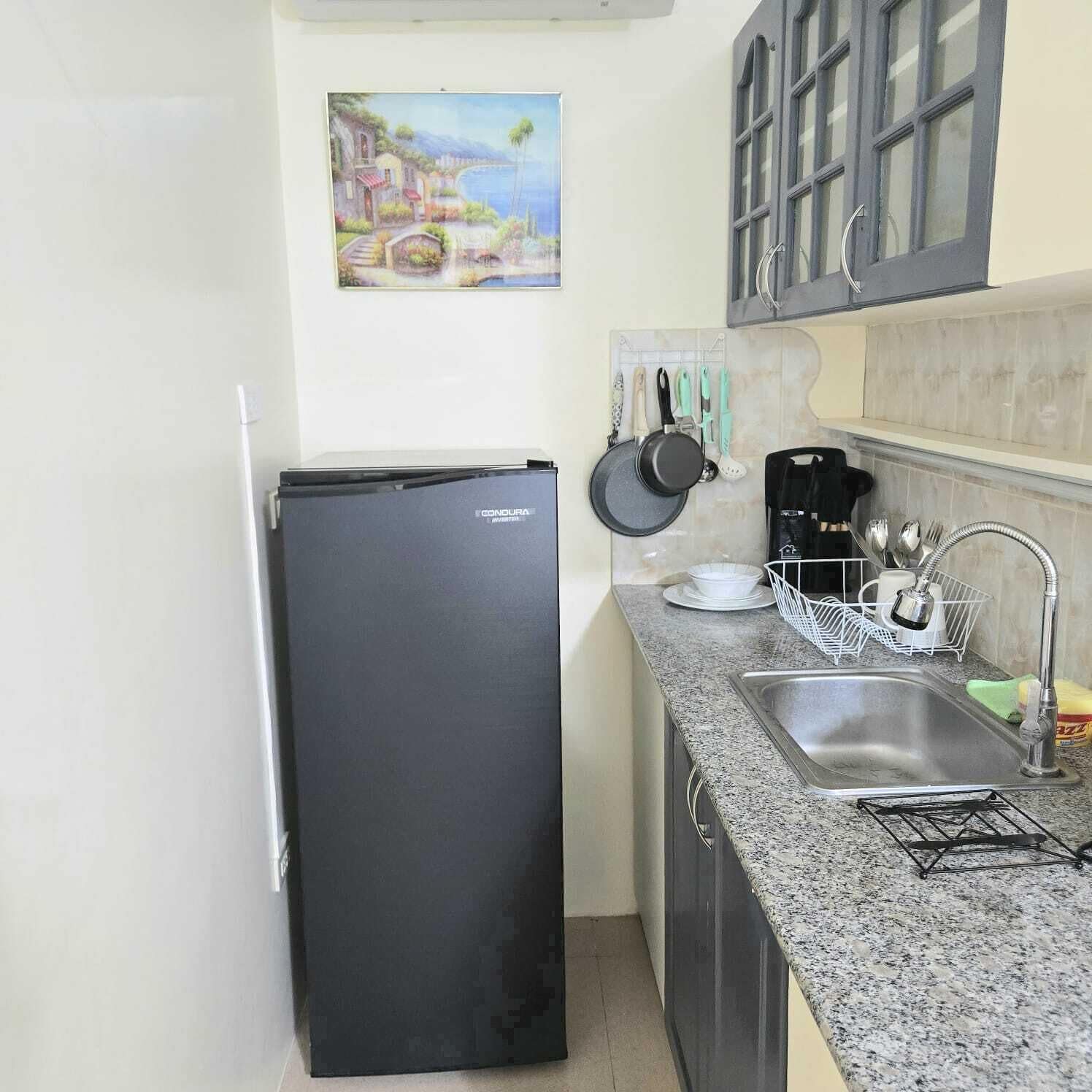 A compact kitchen with a black refrigerator, a granite countertop, a sink with a faucet, and a hanging pot rack.