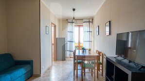 Ein Wohnzimmer mit einer Couch, einem Esstisch mit Stühlen, einem Fernseher und einem Balkon mit Vorhängen.